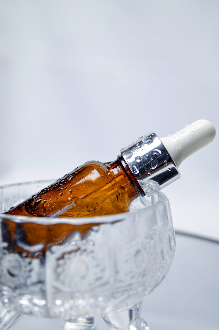 A brown dropper bottle with water droplets rests inside a decorative glass container.